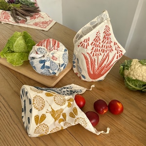 May include: Three reusable fabric bags with different floral and plant patterns. The bags are made of natural cotton and are perfect for storing food or other items. The bags are shown on a wooden table with fresh produce.