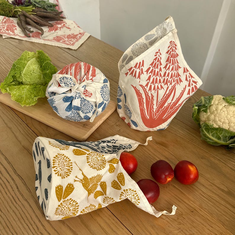 May include: Three reusable fabric bags with different floral and plant patterns. The bags are made of natural cotton and are perfect for storing food or other items. The bags are shown on a wooden table with fresh produce.