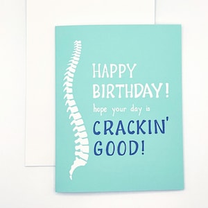 May include: A light blue greeting card featuring a white silhouette of a human spine on the left side. The text reads "HAPPY BIRTHDAY! hope your day is CRACKIN' GOOD!" in white and dark blue lettering. A plain white envelope is positioned behind the card.