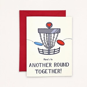May include: A greeting card with a red envelope featuring a disc golf basket illustration with a red heart on top, a blue disc, and a red disc. The text reads: Here's to ANOTHER ROUND TOGETHER! The design uses clean lines and a simple color palette.