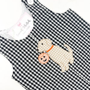 May include: Black and white gingham overall romper with a beige embroidered dog holding a pumpkin. The romper has a white button on the shoulder strap. The label reads "Sweet Shiloh Baby + Child".