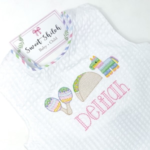 May include: White sleeveless onesie with a pink embroidered name "Delilah" and colorful embroidered images of a taco, a pinata, and maracas. The onesie has a multi-colored trim around the neckline.