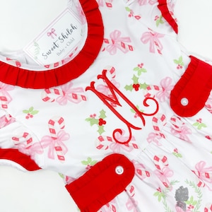 May include: White dress with a red trim and a candy cane and bow print. The dress has a red pocket with a button and a monogrammed "M" on the front. The dress has a label that says "Sweet Shiloh Baby + Child".