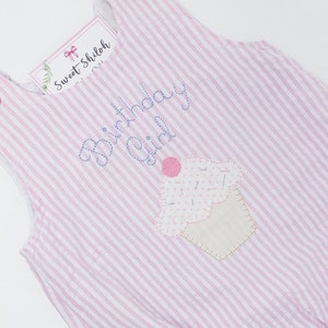 May include: Pink and white striped romper with a cupcake applique and the words "Birthday Girl" embroidered in blue. The romper has button closures on the straps and a tag that says "Sweet Shiloh".
