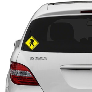 May include: A white car with a yellow warning sign sticker on the back window. The sign has a black silhouette of a Bigfoot walking across the road.