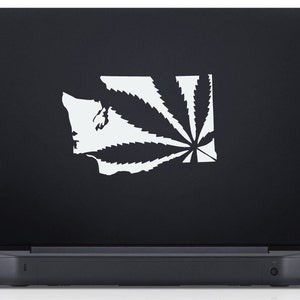 May include: A white vinyl sticker of a marijuana leaf superimposed over the outline of the state of Washington. The sticker is on a black laptop.