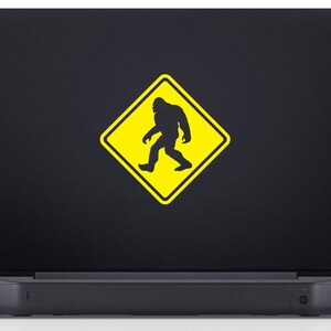 May include: A black laptop with a yellow warning sign sticker featuring a silhouette of Bigfoot walking. The sign reads "Caution Bigfoot Crossing".