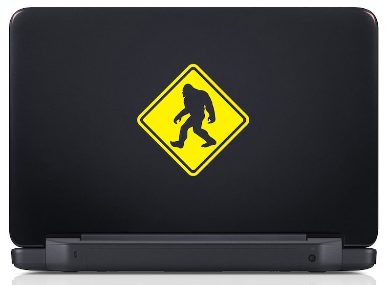 May include: A black laptop with a yellow warning sign sticker featuring a silhouette of Bigfoot walking. The sign reads "Caution Bigfoot Crossing".