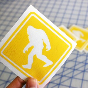 May include: A yellow square sticker with a white silhouette of Bigfoot walking. The sticker is on a white background with a blue grid pattern.