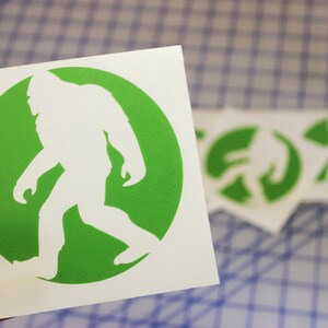 May include: A white vinyl sticker with a green circle and a white silhouette of Bigfoot walking inside the circle.