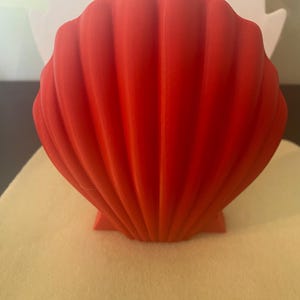 May include: A vibrant red seashell-shaped decorative object. The shell has a ribbed texture and a flat base, sitting on a light beige surface. The background includes a white cutout shape.