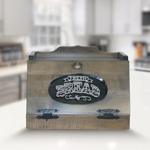 May include: Wooden bread box with a distressed finish. The front features a black oval sign with the words "FRESH BREAD" in white lettering. The box has a hinged door with decorative metal hardware and a small knob.