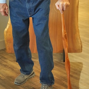 May include: A wooden walking cane with a twisted design. The cane is a light brown color and has a black rubber tip. The person in the image is wearing blue jeans, a light blue shirt, and gray sneakers.
