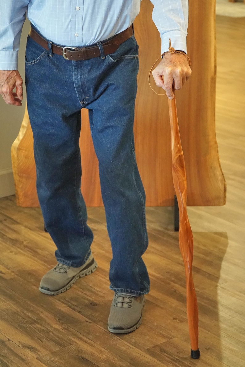 May include: A wooden walking cane with a twisted design. The cane is a light brown color and has a black rubber tip. The person in the image is wearing blue jeans, a light blue shirt, and gray sneakers.