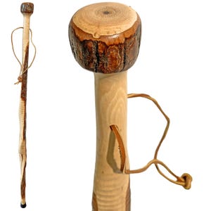 May include: A handcrafted wooden walking stick with a rounded top and a natural wood grain finish. The handle features a leather strap for carrying. The stick is made of light and dark brown wood, with a black rubber tip.