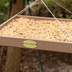 May include: A square, brown bird feeder filled with birdseed, suspended by white cords. The feeder has a simple design with a raised edge and a small, oval label that reads "dutchcrafters". The feeder is outdoors, with a blurred background of trees.