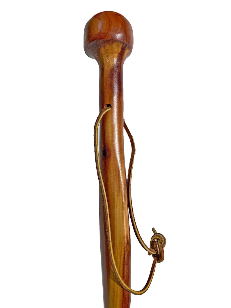 May include: A wooden walking cane with a rounded top and a smooth, polished finish. The cane is a warm brown color with natural wood grain visible. A leather strap is attached near the top. The cane is designed for support and mobility.