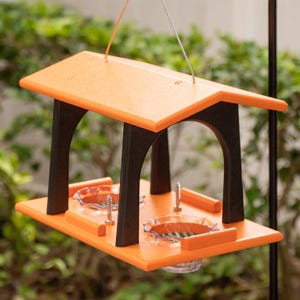 May include: Orange and black bird feeder with two clear feeding dishes. The feeder has a roof and is suspended from a wire.