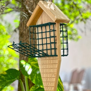 May include: A wooden bird feeder with a green wire mesh insert. The feeder is shaped like a house with a pointed roof and a long, narrow base. It is hanging from a hook.