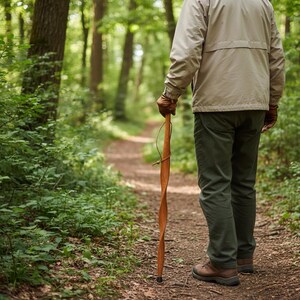 May include: A person walking on a forest path, holding a wooden walking stick. The stick is a light brown color with a twisted design. The person is wearing a tan jacket, green pants, and brown boots. The path is surrounded by green foliage.