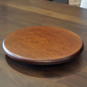 May include: A round, wooden serving tray with a rich, reddish-brown finish. The tray has a smooth surface and a slightly raised edge, perfect for displaying food or decor. The wood grain is visible.