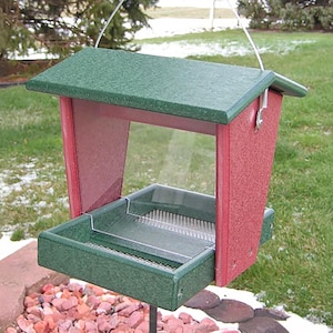 May include: A red and green bird feeder with a clear plastic shield and a metal hanging hook. The feeder has a green roof and a green tray with a wire mesh bottom.