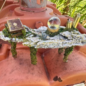 May include: A miniature fantasy scene featuring a small book, a crystal ball, and other decorative elements on a moss-covered platform. The platform is supported by moss-covered pillars. The scene is set against a rusty orange background.