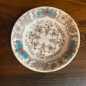 May include: A round, off-white plate with a detailed floral pattern in brown, teal, and orange. The plate features a central floral design and a decorative border. The plate is set on a dark wooden surface.