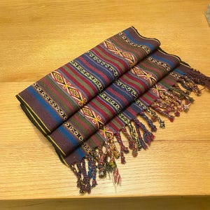 May include: Folded textile scarf with a colorful geometric pattern in red, blue, green, and yellow. The scarf has a fringed edge and is made of woven fabric. The scarf is folded on a wooden surface.