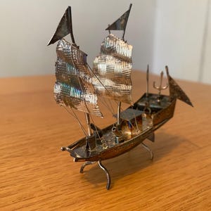 May include: A miniature silver-toned metal junk ship model featuring two textured sails, a pointed bow, and a small trident-shaped ornament on the deck. The vessel is supported by four thin, curved metal legs that elevate the hull above the surface.