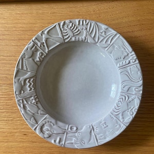 May include: A round, light gray ceramic bowl featuring a wide, embossed rim decorated with twelve distinct zodiac symbols. The center of the bowl is smooth and concave, while the outer border displays raised relief carvings of astrological signs.