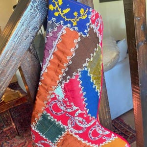 May include: A colorful patchwork Christmas stocking with a variety of fabric pieces in red, blue, orange, and brown. The stocking is decorated with white stitching and a blue top with yellow floral embroidery. A festive holiday decoration.