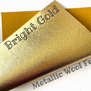 May include: A close-up of a metallic gold wool felt sheet. The sheet is textured and has a subtle sheen. The text "Bright Gold Metallic Wool Felt" is printed on the sheet.