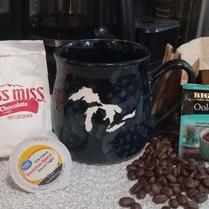 May include: A dark blue mug with a white graphic of a lake, next to a packet of Swiss Miss hot cocoa mix, a coffee pod, a tea bag, and coffee beans. The tea bag is labeled "Oolong Tea".