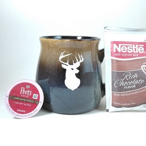 May include: A brown and blue ceramic mug with a white deer silhouette, a Peet's Coffee Holiday Blend K-Cup, and a Nestle Hot Cocoa Mix packet. The cocoa mix is labeled "Rich Chocolate Flavor" and is 99.9% caffeine-free.
