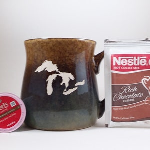 May include: A brown and green ceramic mug with a white outline of the Great Lakes.  The mug is next to a packet of Nestle Rich Chocolate Hot Cocoa Mix and a single-serve Peet's coffee pod.