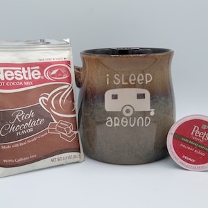 May include: A brown ceramic mug with the text "i sleep around" and a white camper van graphic.  A single-serve coffee pod with the text "Peet's Coffee Dark Roast Coffee Holiday Blend" is in the foreground.  A packet of Nestle hot cocoa mix is in the background.