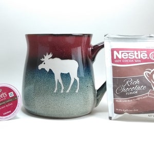 Könnte beinhalten: Eine rot-weiß-blaue Keramiktasse mit einem Elch-Motiv. Die Tasse steht neben einer Packung Nestle Rich Chocolate Hot Cocoa Mix. Die Packung ist rot, weiß und braun mit dem Nestle-Logo und dem Text "Rich Chocolate Flavor".