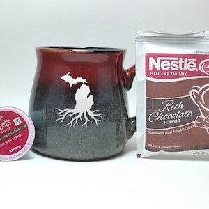 May include: A red and black ceramic mug with a white outline of the state of Michigan and tree roots. A single-serve coffee pod and a packet of Nestle Rich Chocolate Hot Cocoa Mix are also pictured.