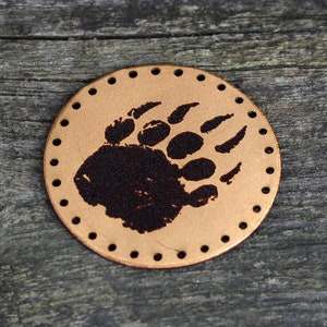 May include: A round leather patch with a brown bear paw print in the center. The patch has a stitched edge with holes around the perimeter.