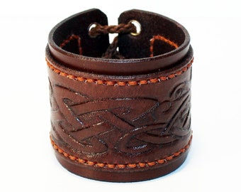 May include: A brown leather bracelet with an intricate Celtic knot design. The bracelet has an adjustable strap and is accented with orange stitching.