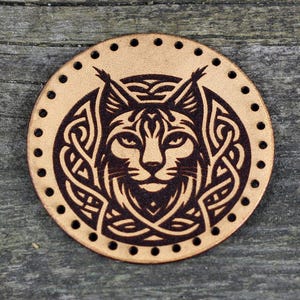 Könnte beinhalten: Runde, hellbraune Lederscheibe mit detailliertem, dunkelbraunem Luchsgesicht-Design. Der Luchs ist von einem komplizierten keltischen Knotenmuster umgeben. Der Rand hat gleichmäßig verteilte Löcher zum Nähen. Auf einem verwitterten Holzgrund.