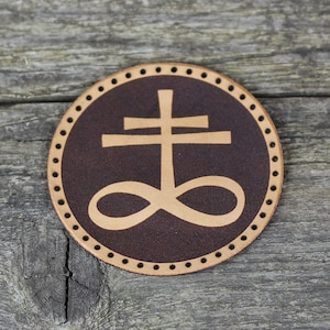 Peut inclure: Un dessous de verre en cuir marron avec un symbole en cuir beige d'une croix surmontée d'un symbole d'infini. Le dessous de verre a un bord cousu décoratif.