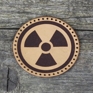 May include: A brown leather patch with a black radiation symbol in the center. The patch has a stitched border.