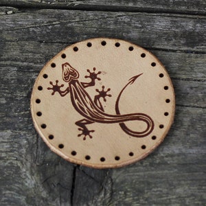 Pode incluir: Um remendo redondo de couro marrom com um design de lagarto marrom. O remendo tem uma borda costurada com pequenos furos ao redor do perímetro.