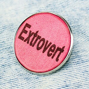 Op de afbeelding: Een ronde, roze pin met het woord "Extrovert" in donkerbruine letters. De pin heeft een zilverkleurige metalen rand en is geplaatst tegen een lichtblauwe denim achtergrond.