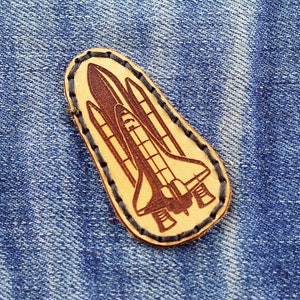 Op de afbeelding: Een bruine leren patch met een gestikte rand, met een gedetailleerde illustratie van een spaceshuttle in de vlucht.