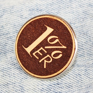 Può includere: Un bottone rotondo marrone con bordo argentato. Il bottone presenta il numero "1" e le lettere "ER" in un colore più chiaro, in contrasto con lo sfondo marrone scuro. È appoggiato su una superficie di denim blu.