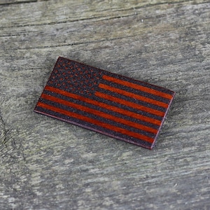 Puede incluir: Una pequeña bandera americana de madera con rayas rojas y blancas y estrellas azules. La bandera está hecha de madera oscura y tiene un aspecto desgastado.