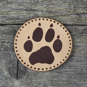 Może przedstawiać: Brązowa skórzana łatka z brązowym nadrukiem łapy. Łatka ma obszyty brzeg.